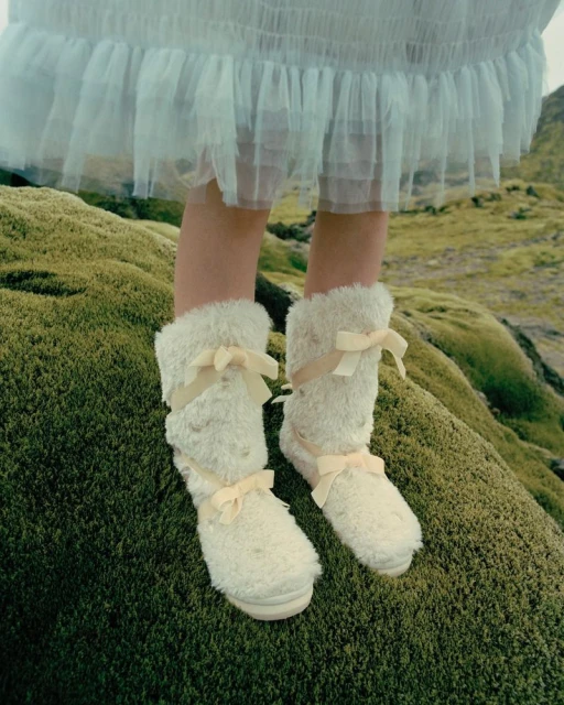 Close-up of a person wearing fluffy, cream-colored, mid-calf boots with light yellow bows standing on mossy ground outdoors.