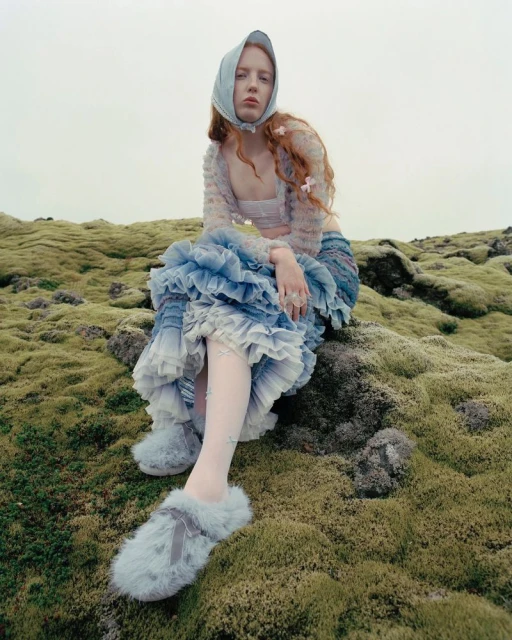 A red-haired young woman in a blue tulle dress and headscarf sits on a mossy hill in an outdoor setting, fashion-forward and whimsical.