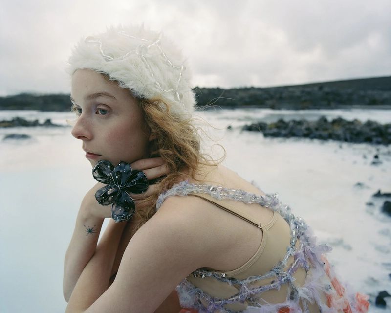 Young woman poses in a light pink and beige dress with a dark flower accessory, wearing a white hat, in a pale blue lagoon landscape.