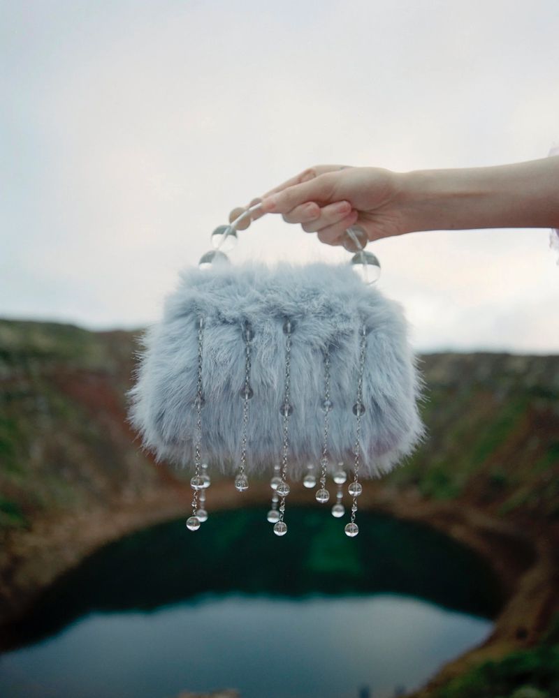 A furry blue purse with dangling crystals is held by a hand in front of a crater lake during daytime, showcasing fashion accessory.