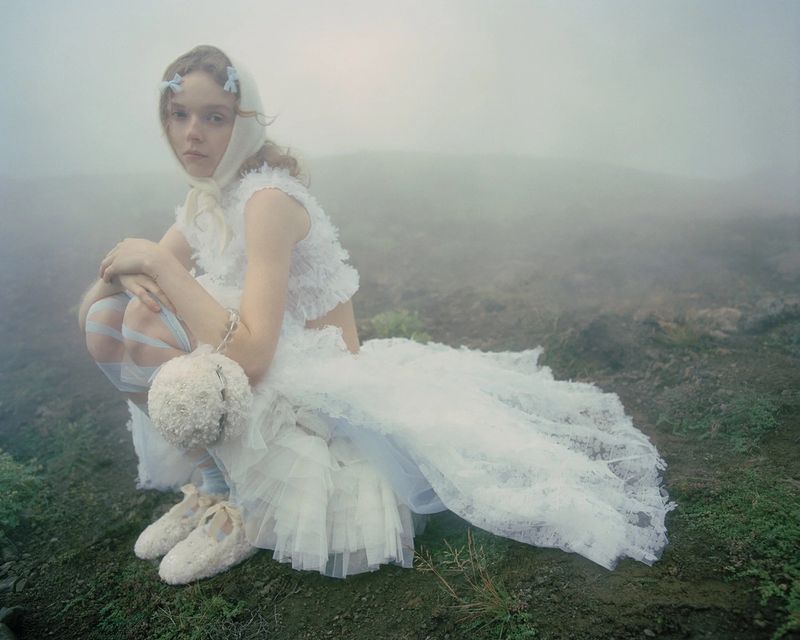 Young woman in a white dress and headscarf crouching in a foggy landscape, holding a ball and wearing ballet flats.