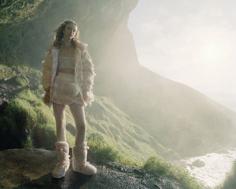 Young woman in matching pastel fringed jacket and skirt, plus fuzzy boots poses on a rocky outcrop with mountain views and water.