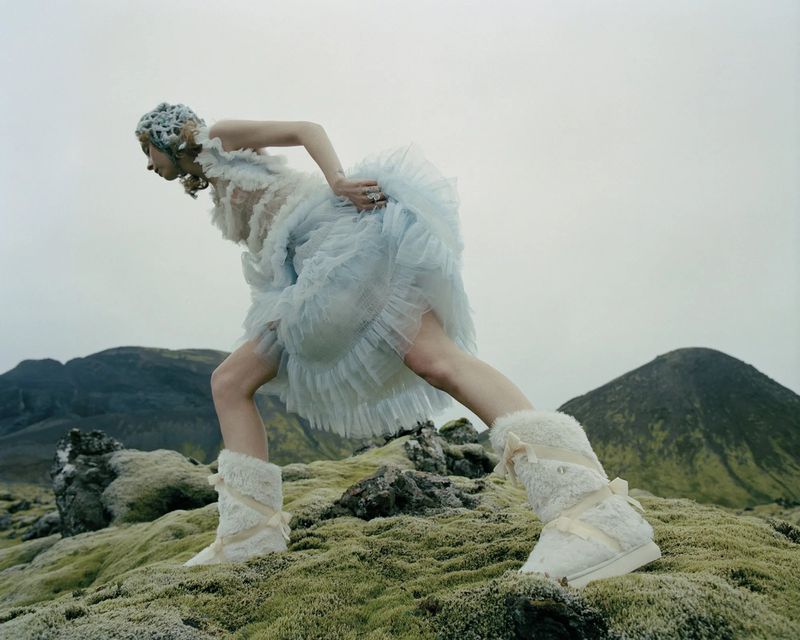 A woman in a blue dress and knit hat kneels on mossy ground in white boots, with a backdrop of misty mountains on an overcast day.