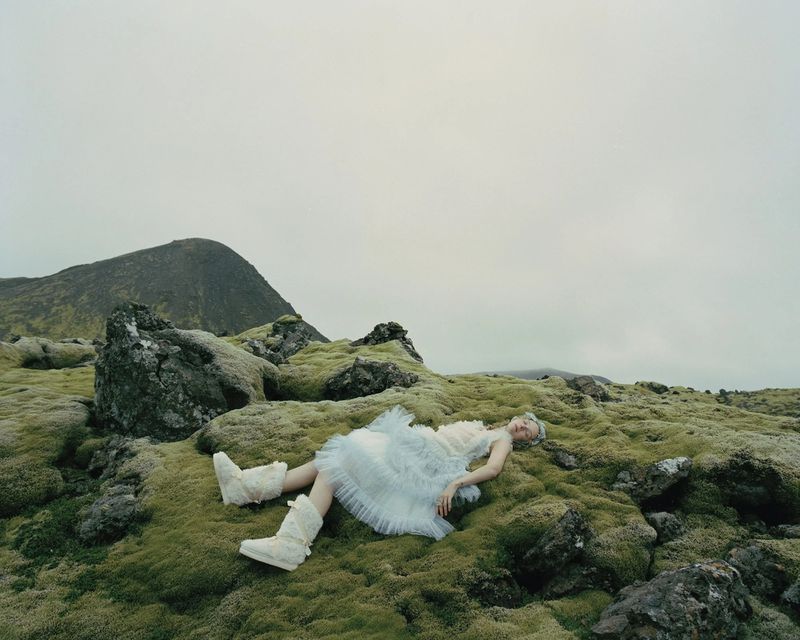 Fashion shot of a woman lying on mossy ground in a light blue dress and furry boots, with a mountain in the background under an overcast sky.