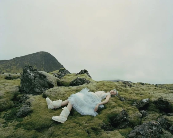 Fashion shot of a woman lying on mossy ground in a light blue dress and furry boots, with a mountain in the background under an overcast sky.