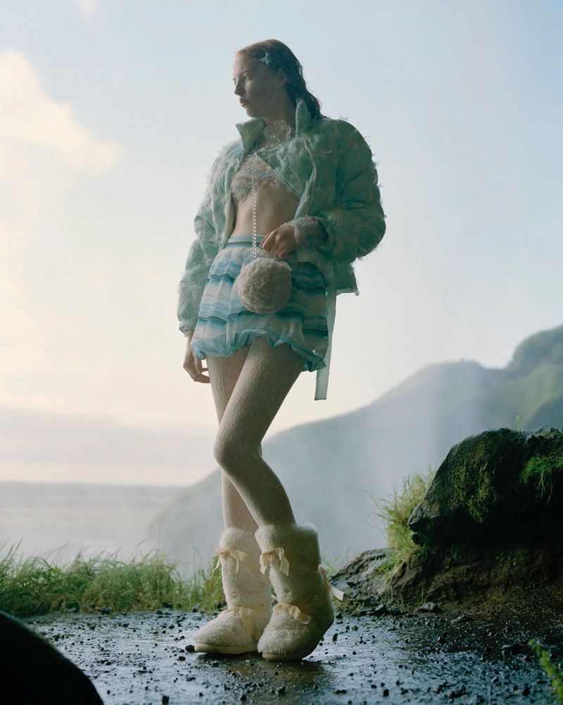 A woman in fashionable outerwear and furry boots stands on a rock near a waterfall against a bright, overcast sky in an outdoor setting.