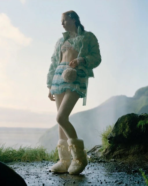 A woman in fashionable outerwear and furry boots stands on a rock near a waterfall against a bright, overcast sky in an outdoor setting.
