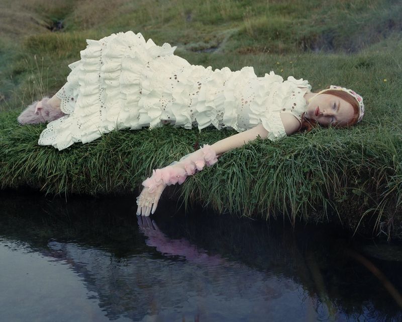 Red-haired woman in ruffled white dress lies on grassy riverbank, arm dangling in water, wearing bonnet and lace gloves, eyes closed.