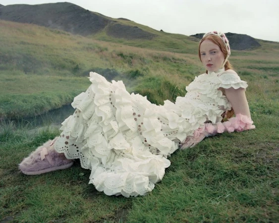 Red-haired female model in a ruffled white dress lying on a grassy hill wearing pink fuzzy footwear.