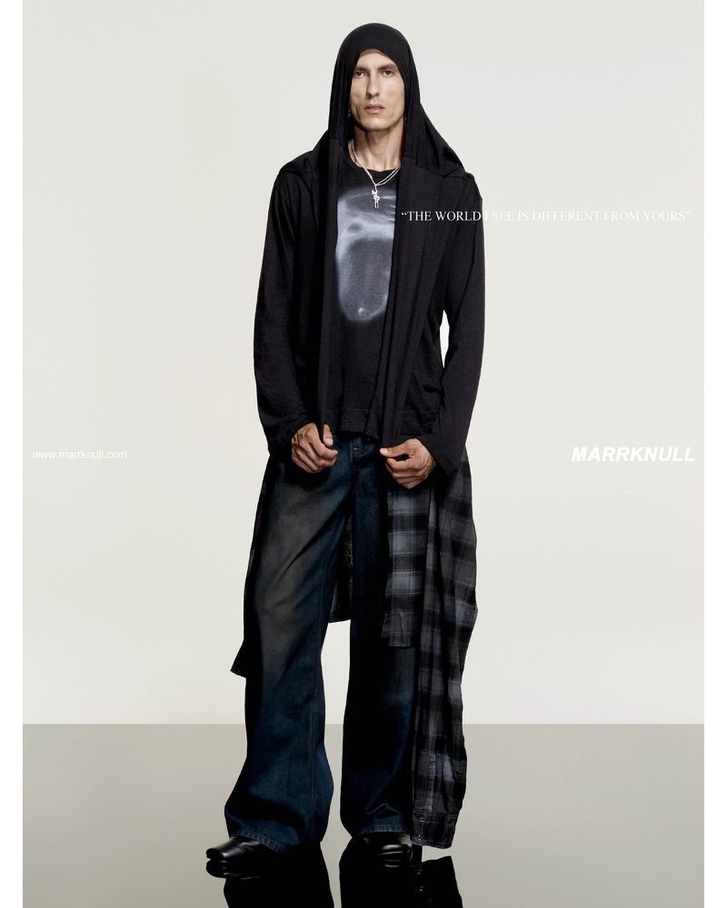 Fashion portrait of adult male wearing dark hooded jacket and wide-leg jeans, standing in a studio with a reflective surface.