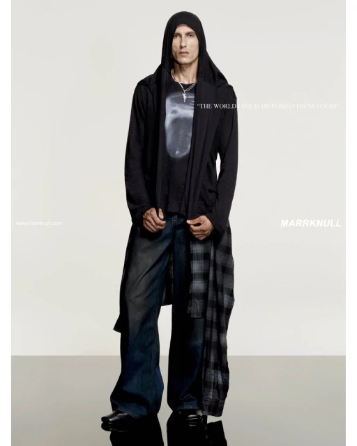 Fashion portrait of adult male wearing dark hooded jacket and wide-leg jeans, standing in a studio with a reflective surface.