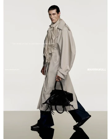 A stylish adult male model strides confidently, wearing a tan trench coat and carrying a black fringed handbag in a studio setting.