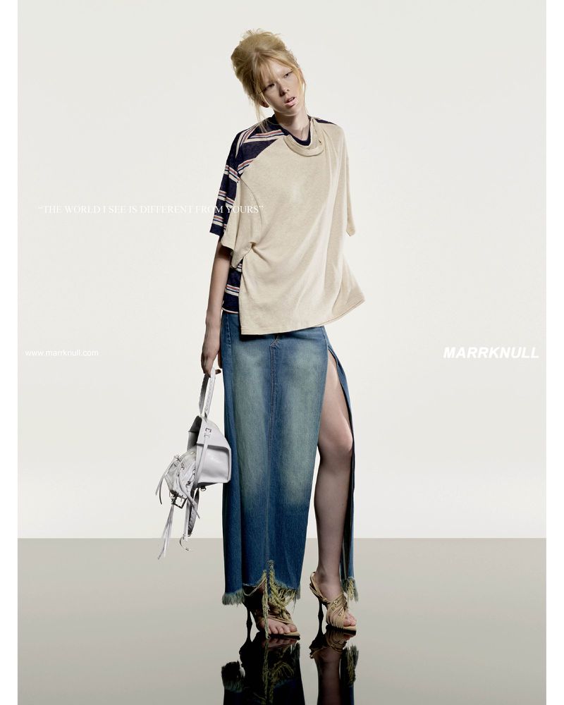 Fashion editorial shot of a blonde model in a beige top and long denim skirt holding a white purse against a studio backdrop.