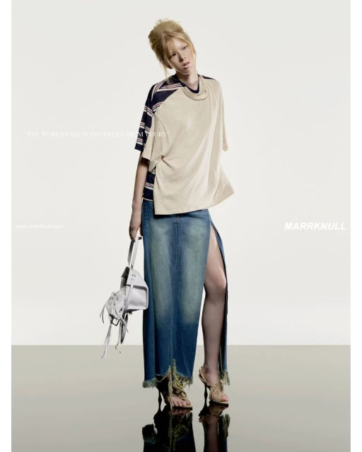 Fashion editorial shot of a blonde model in a beige top and long denim skirt holding a white purse against a studio backdrop.