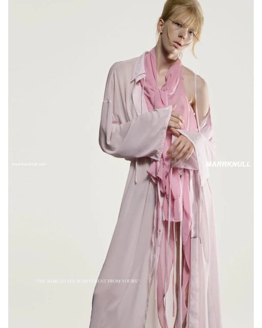 A slender fashion model poses in a flowing light-pink ensemble with an asymmetrical draped scarf against a pale backdrop.
