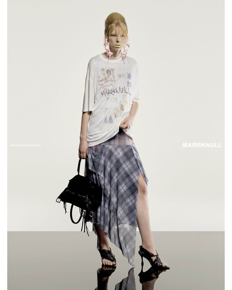 Fashion shoot of a blonde woman wearing a graphic t-shirt, plaid skirt, and black heels, holding a purse against a white backdrop.