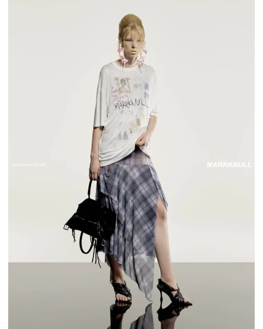Fashion shoot of a blonde woman wearing a graphic t-shirt, plaid skirt, and black heels, holding a purse against a white backdrop.