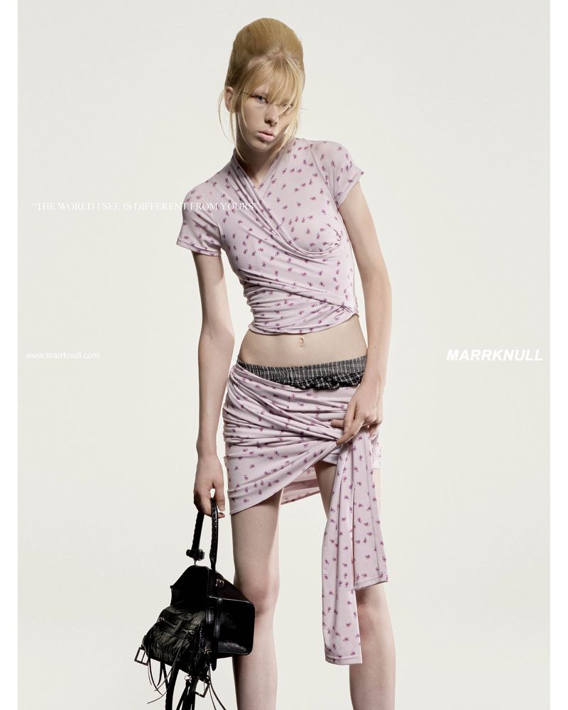 A young, slender fashion model in a printed light purple wrap top and skirt is holding a black leather fringe bag in a studio shot.