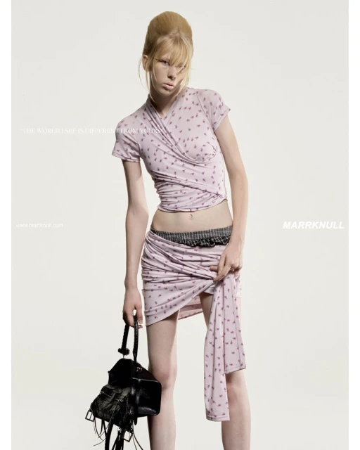 A young, slender fashion model in a printed light purple wrap top and skirt is holding a black leather fringe bag in a studio shot.