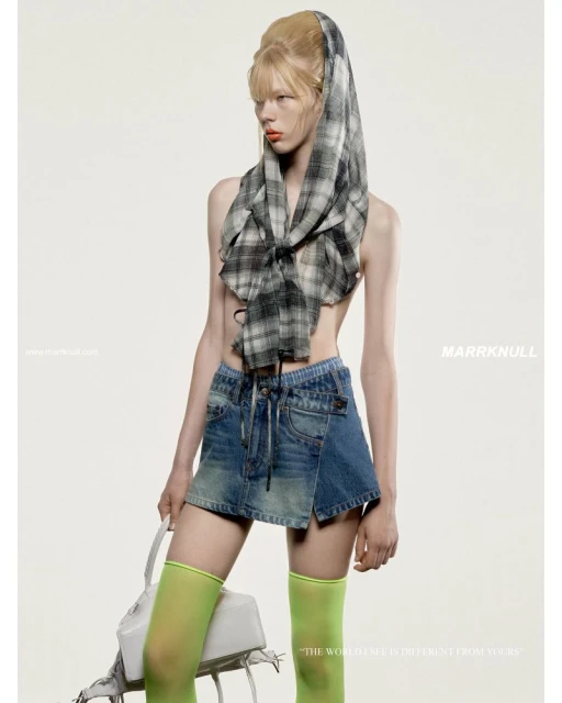 Fashion shot of a fair-skinned adult female model in a denim miniskirt, with a plaid scarf head covering, translucent knee socks, and a white purse.