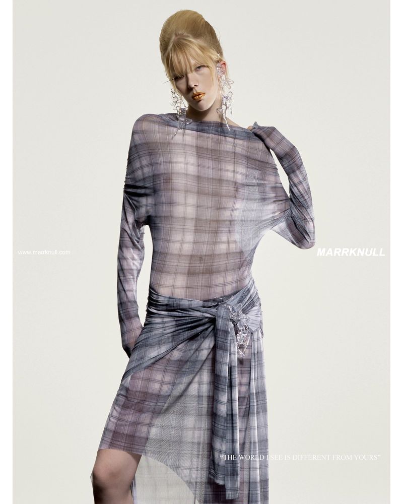 Fashion editorial shot of a blond woman in a sheer plaid dress with statement earrings, standing in front of a pale neutral background.