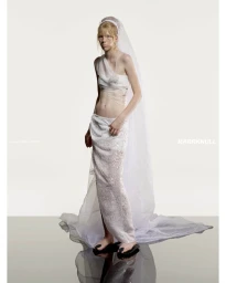 Model in a sheer white layered skirt and veil standing against a light grey studio background with a reflective floor.