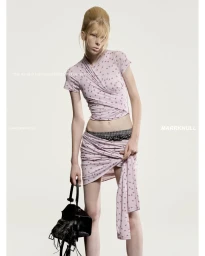 Model in printed pink top and skirt holding black bag against plain white background