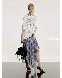 Model in printed white t-shirt and grey plaid skirt holding black handbag against a white background