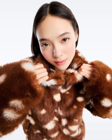 A woman with dark hair poses in a brown faux fur coat with white spots, in a bright studio setting, looking directly at the camera.