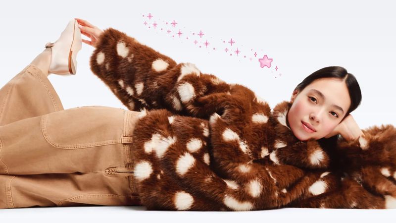 Woman lounges in a brown and white faux fur coat and tan pants, one leg raised showing a cream-colored flat shoe. Soft, diffused studio light.