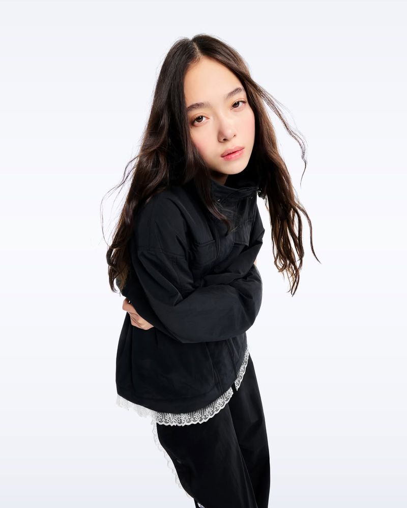 Portrait of a young person with long, dark hair wearing a black jacket and dark pants, posing against a white studio backdrop.