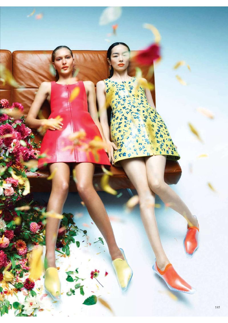 Two women in vibrant mini dresses pose on a tan couch surrounded by colorful flowers and falling petals in a studio setting.
