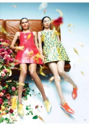Two female models in colorful dresses seated on a brown leather sofa with scattered flowers and floating petals against a light blue backdrop.