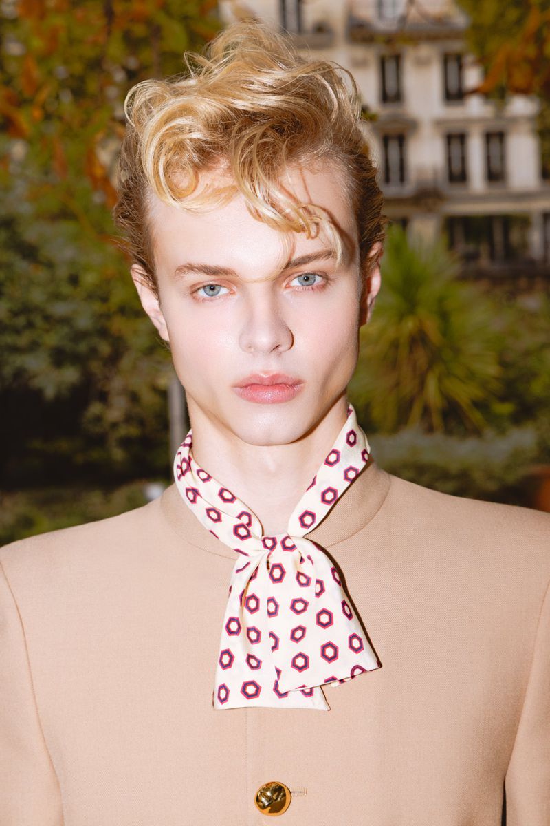 Portrait of a young male model with styled blond hair wearing a tan jacket and a patterned neck scarf outdoors.