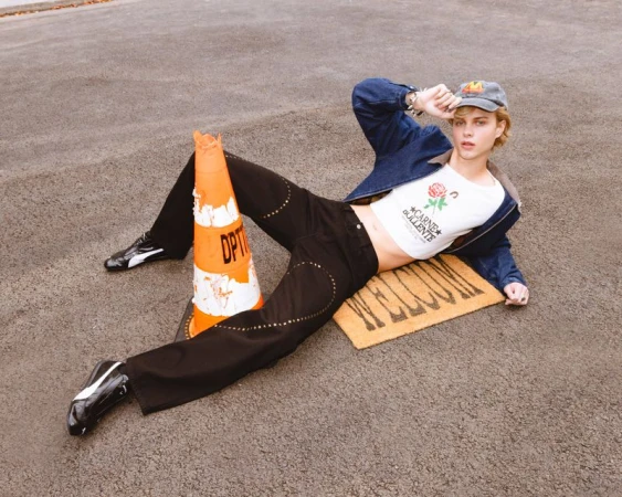 Man in denim jacket lies on welcome mat next to traffic cone in urban setting, wearing a cropped T-shirt and jeans with embellishments.