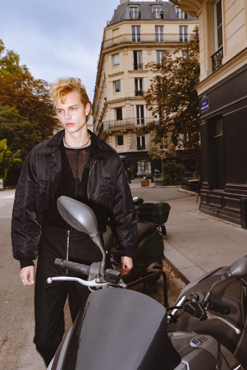 A blonde male fashion model poses on a city street next to a scooter, wearing a black jacket and pants in an outdoor setting.