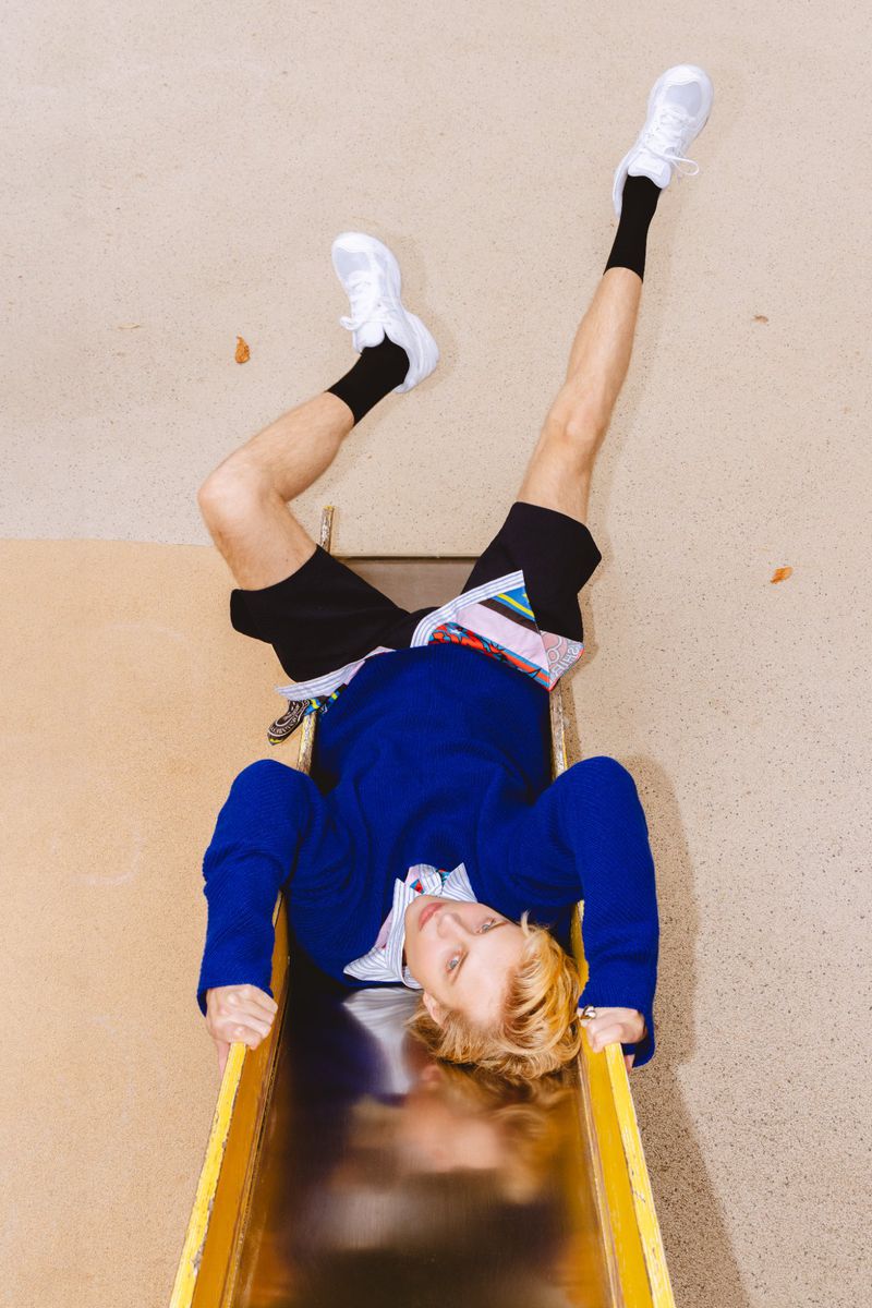 Young adult man slides down a yellow playground slide. He wears a blue jacket, black shorts, white socks and sneakers, and has blond hair.