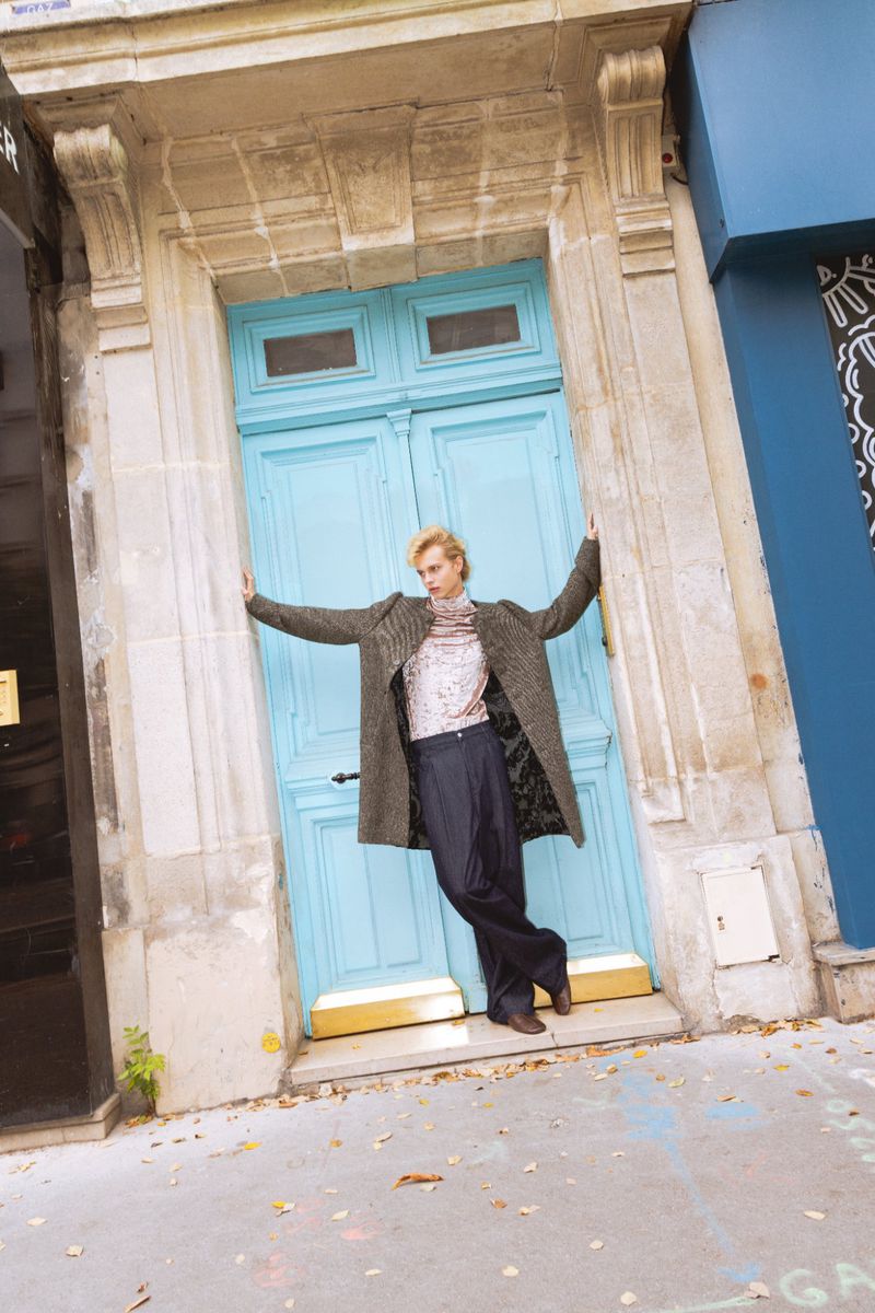 Fashionable woman poses in a grey coat, sequined top, and black pants in front of a light blue door on a city street.