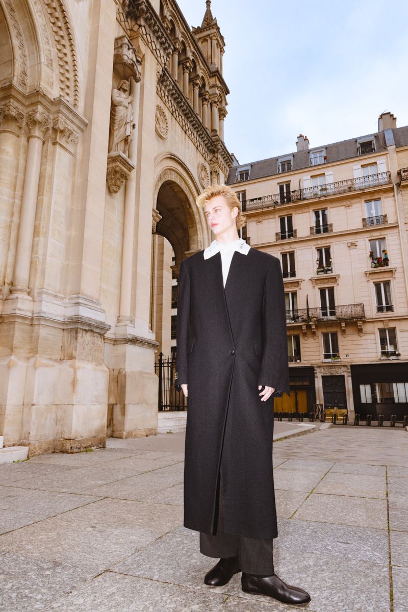 A blonde man in a long black coat poses on a stone plaza with an ornate stone building and residential buildings in the background.
