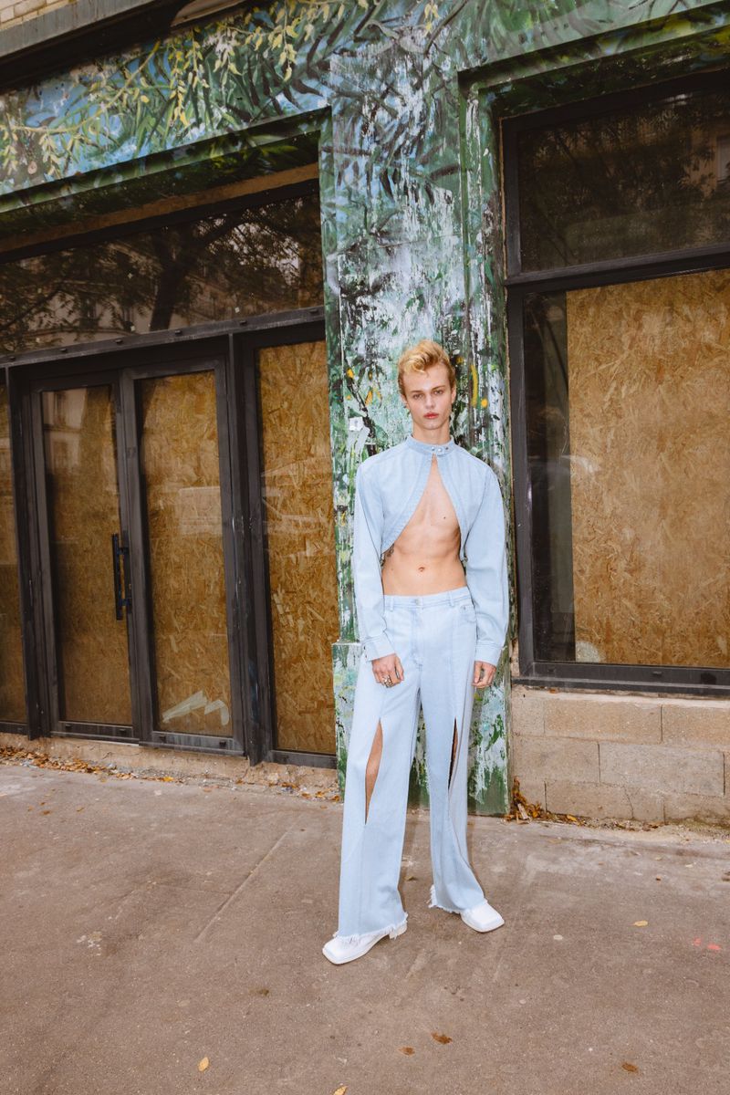 Androgynous person in pale blue outfit poses against an old building with boarded-up windows and a mural of leafy vegetation.