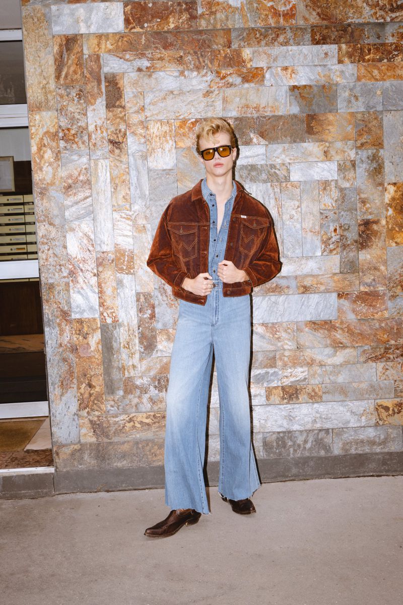 Stylish man in denim and corduroy posing against a stone wall. He wears sunglasses and brown shoes in an outdoor setting.