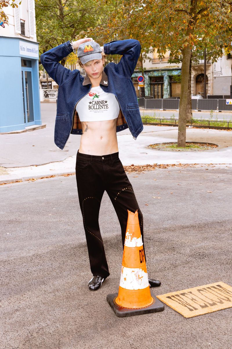 Person in crop top and jeans poses with traffic cone and welcome mat outdoors on city street in a fashion editorial shot.