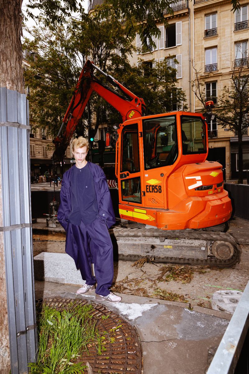 Young adult poses in front of an orange excavator in an urban setting with trees and buildings in the background, street-style fashion.