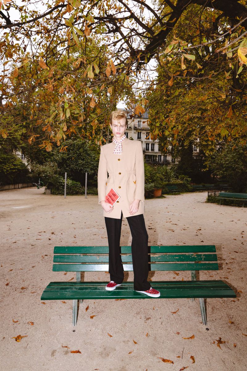 Blonde adult stands on a green park bench wearing a beige jacket and black pants, surrounded by fall foliage in an urban park setting.