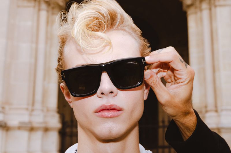 A person with blond hair wearing black sunglasses and a white shirt is positioned indoors against a backdrop of ornate columns.