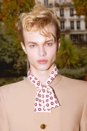 Portrait of a young male model with styled blond hair wearing a tan jacket and a patterned neck scarf outdoors.