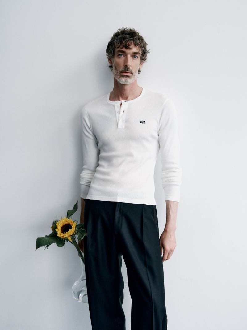 Adult man stands indoors holding a sunflower, wearing a white long-sleeve shirt and black pants against a white wall in a portrait shot.