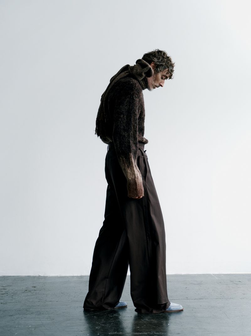 Fashionable adult male with tousled hair wearing a brown sweater and pants in a studio setting with a white background.