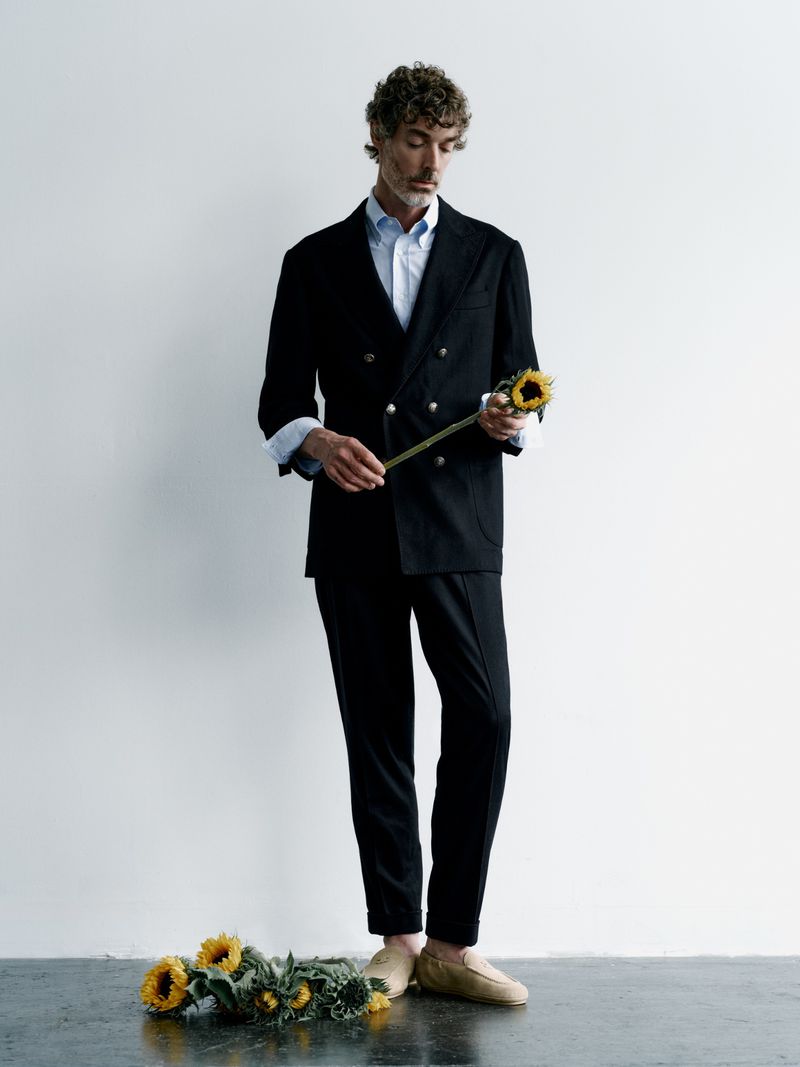 Adult man in a suit standing indoors holding a sunflower, with a group of sunflowers lying at his feet on a grey surface.