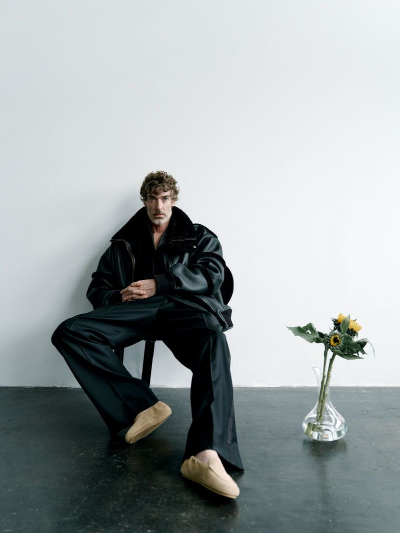 Stylish man in oversized jacket poses indoors while seated. Features sunflower arrangement, dark monochromatic tones, and studio lighting.